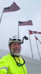 A man wearing a white bike helmet, neon yellow jersey, and sunglasses around his neck with American and Canadian flags waving behind him.
