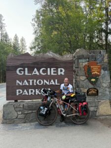 A photo of a man and his bicycle in front of the Glacier National Park sign with evergreen trees in the background.