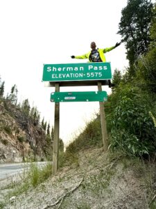 A man in a neon yellow windbreaker posing on top of a road sign that reads Sherman Pass, Elevation 5,575.