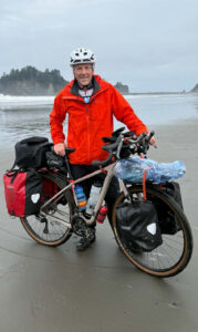 A man in a red windbreaker with his packed bicycle on a beach on a rainy day.