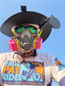 A man wearing a respirator, hat and sunglasses, along with a Ride Pat Ride cycling jersey.