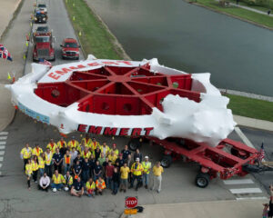 A giant piece of technology on a tractor trailer, along with a large group of people, taken from an aerial view.