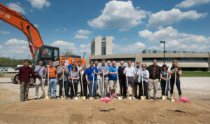 A group of people with shovels at a construction site with equipment and buildings behind them.