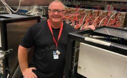 A man wearing a lanyard and smiling, surrounded by equipment for physics research.
