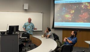 A man standing in a classroom with two professors seated at a table and a presentation on a screen.