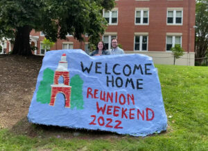 A photo of a couple standing behind the UMW Spirit Rock painted blue with an image of the Bell Tower and the words 'Welcome Home Reunion Weekend 2022.'
