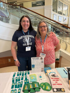 Two people, one wearing a Mary Washington College shirt and the other wearing a red/pink print shirt, with both wearing lanyards, smiling and standing in the lobby of the Cedric Rucker University Center. Books and swag are on the table.