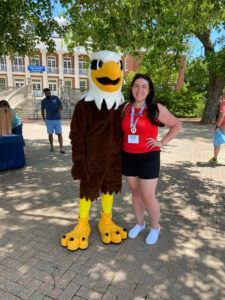 Sammy D. Eagle mascot on the left next to a person with long brown hair, wearing a summery red tank top and black shorts, a lanyard, and white sneakers in front of UMW's Lee Hall.