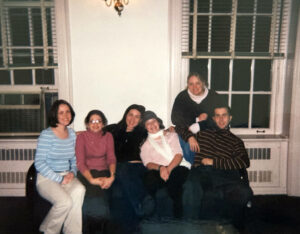 A group of six students sitting on a couch against a white wall in between two windows in a 2000s photo.