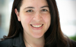 A photo of a smiling person with long, dark brown hair and a black suit jacket and shirt with a silver medallion necklace.