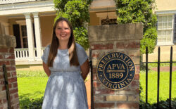 A student wearing a summery dress, smiling and standing next to a brick column with a round plaque next to an iron fence. A historic building is in the background.