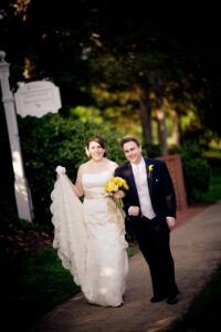 A couple in a wedding gown and tux, where the bride is holding yellow flowers. In the background is a sidewalk, brick wall and a sign for the Jepson Alumni Executive Center.