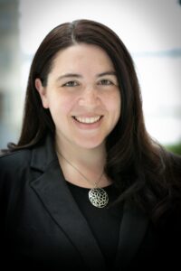 A photo of a smiling person with long, dark brown hair and a black suit jacket and shirt with a silver medallion necklace.
